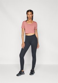 Rosa kortärmad t-shirt med Nike-logga, tillsammans med svarta högmidjade leggings. Modell som bär svarta träningsskor, står framför en neutral bakgrund.