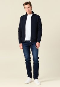 Giacca zip-up blu navy con colletto alto, con cuciture bianche a contrasto, abbinata a jeans blu scuro aderenti e sneaker bianche.