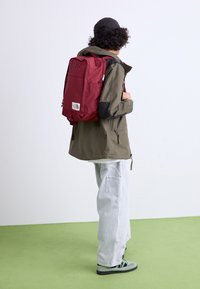Rød ryggsekk med glidelåser, logo-patch og polstret rygg. Brukt over en olivenjakke, kombinert med lyse jeans og grønne sko.