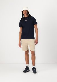 Marineblå t-skjorte med kort erme og hvitt logo, beige shorts, hvit bøttehatt og marineblå sneakers med teksturert design og synlige snørebånd.