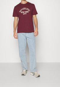 Camiseta de algodón burdeos con "GAP ATHLETIC NEW YORK CITY" impreso en crema. Llevada con jeans de color azul claro y zapatillas beige.