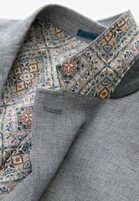 Sivi tkanjeni blazer s uzorkom bež i plave unutrašnjosti. Značajne detalje uključuju teksturu riblje kosti i složeni geometrijski uzorak podstave.
