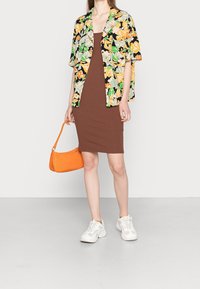Camicia a maniche corte floreale con motivi verdi, gialli e arancioni, abbinata a un vestito aderente marrone, borsa arancione e sneaker bianche.