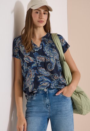 Jeune femme portant un chemisier bleu à motif cachemire, un jean bleu, une casquette beige et portant un sac matelassé vert clair sur son épaule.