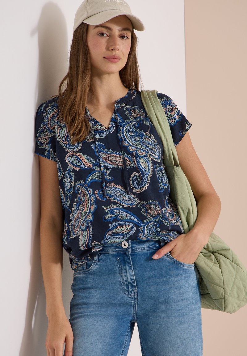 Jeune femme portant un chemisier bleu à motif cachemire, un jean bleu, une casquette beige et portant un sac matelassé vert clair sur son épaule.