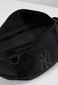 Sac à taille en nylon noir au design horizontal, avec des compartiments zippés et un grand logo en relief. Intérieur doublé et texturé.
