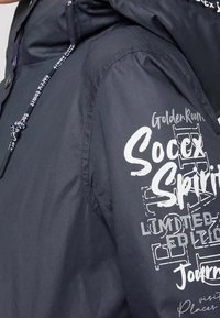 Ciemnoniebieski rękaw kurtki z białym nadrukiem, w tym "Złota Sala", "Duch Piłki Nożnej" oraz "Edycja Limitowana", z kapturem i sznurkiem do regulacji.