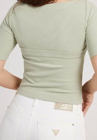 Mujer vistiendo una blusa de manga corta de canalé verde claro y unos jeans blancos de cintura alta con bolsillos traseros y etiqueta de marca.