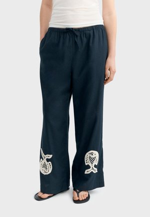 Pantaloni larghi blu navy con disegni bianchi a uncinetto di frutta sulle gambe inferiori, indossati con infradito nere e una maglia bianca infilata dentro.