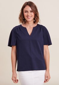 Top blu navy a maniche corte con scollatura a V, caratterizzato da arricciature sulle spalle e un tessuto testurizzato. Abbinato a pantaloni bianchi.