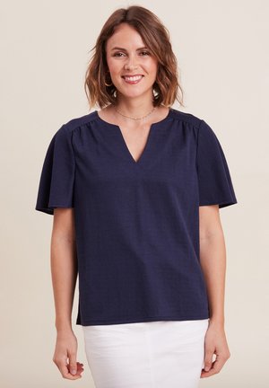 Haut bleu marine à manches courtes avec un col en V, présentant des fronces aux épaules et un tissu texturé. Associé à un pantalon blanc.