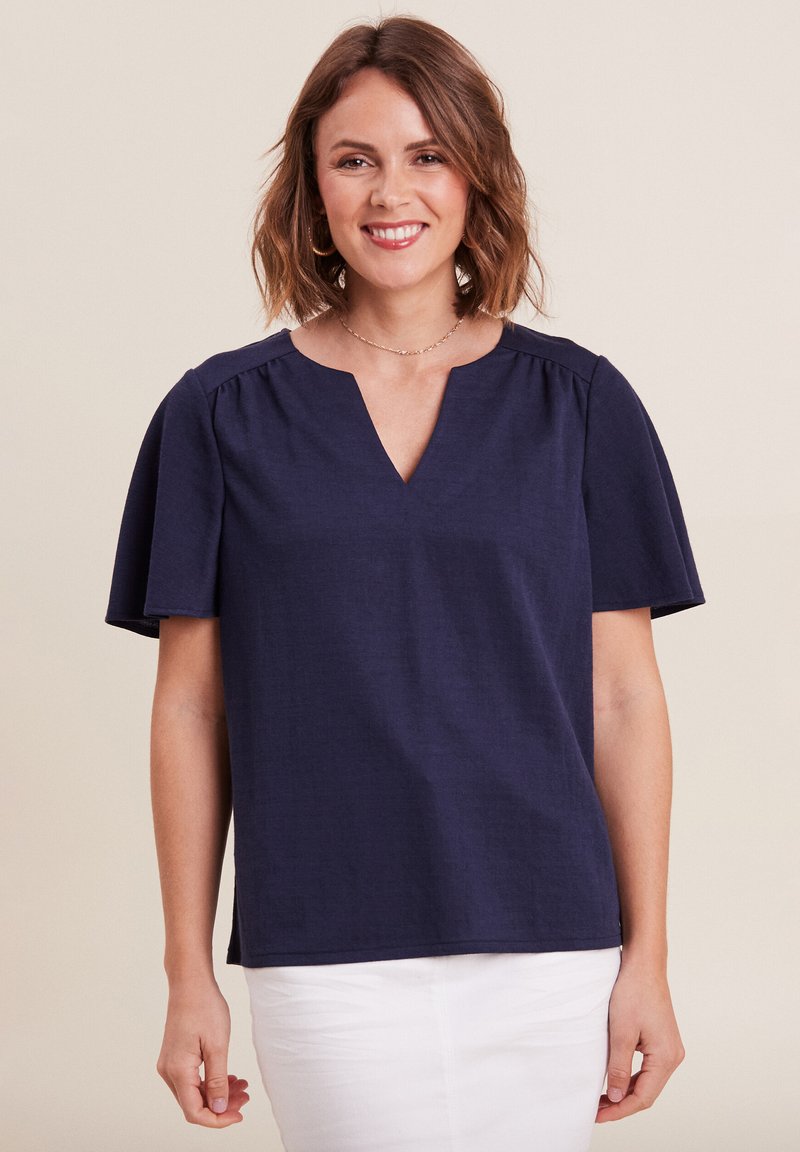 Top blu navy a maniche corte con scollatura a V, caratterizzato da arricciature sulle spalle e un tessuto testurizzato. Abbinato a pantaloni bianchi.