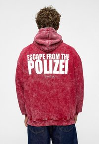 Rode, oversized hoodie van een gestructureerde, gewassen stof. Bevat grote witte tekst "ESCAPE FROM THE POLIZEI" op de achterkant. Met een capuchon.