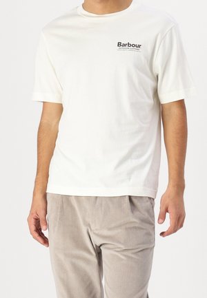 Man die een wit Barbour T-shirt met korte mouwen en lichtbeige ribfluwelen broek draagt, staand tegen een effen achtergrond.