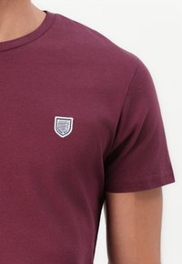 Bordo pamučna majica s okruglim izrezom i kratkim rukavima. Ima mali bijeli izvezeni oznaku koja izgleda kao štit na prsima.