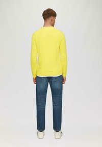 Helles gelbes Langarmshirt mit einem runden Ausschnitt, kombiniert mit blauen Straight-Leg-Jeans und weißen Sneakern. Ansicht von hinten.