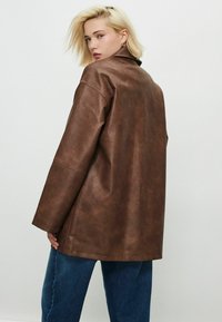 Personne blonde portant une veste en cuir marron ample et un jean bleu, regardant par-dessus son épaule contre un fond clair.