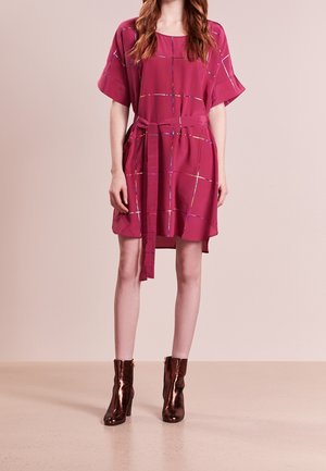 Frau trägt ein kurzärmeliges magentafarbenes Kleid mit Taillenband und dezentem Karomuster, kombiniert mit metallisch bronzenen Knöchelstiefeln.