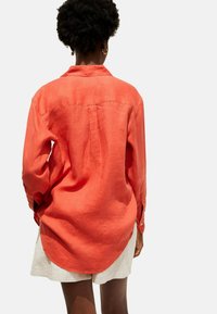 Rotes Leinen-Oversize-Hemd mit langen Ärmeln, abgerundetem Saum und geknöpften Manschetten. Der Stoff hat eine natürliche Struktur und einen leichten Glanz.