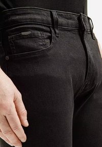 Czarne dżinsy z dopasowanym krojem, z małą kieszonką z przodu, szlufkami na pasek i tonowanymi szwami. Gładka faktura, minimalna ilość elementów metalowych.