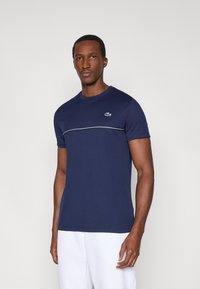 Granatowy bawełniany t-shirt z krótkimi rękawami, okrągłym dekoltem i poziomym białym paskiem na wysokości klatki piersiowej. Po lewej stronie znajduje się zielone logo.