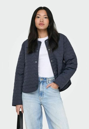 Žena s dlhými tmavými vlasmi oblečená v námorníckej prešitej bunde, bielej košeli a svetlo modrých džínsach, držiaca v jednej ruke čiernu kabelku.