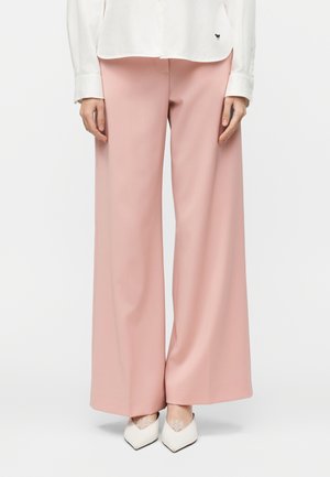 Nő halvány rózsaszín, bőszárú nadrágot, fehér, csipkés, hegyes orrú cipőt és fehér inget visel, amelynek szegélyénél egy kis fekete logó található.