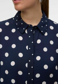 Chemise boutonnée bleu marine avec un tissu texturé, ornée de grands pois blancs et d'un col classique. Les boutons sont de la même couleur que le tissu.