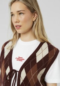 Gilet en argyle tricoté dans des teintes de bordeaux, crème et marron, porté sur un t-shirt blanc avec un texte rouge. Présente un devant noué et une encolure ronde.
