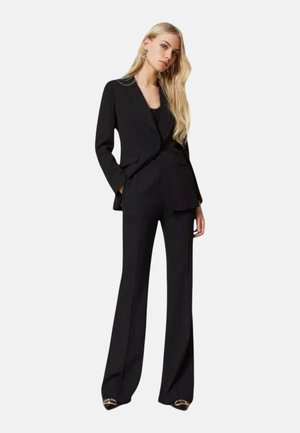 Donna con lunghi capelli biondi che indossa un blazer nero, pantaloni neri ampi e scarpe nere con dettagli dorati, in piedi con le mani nelle tasche.