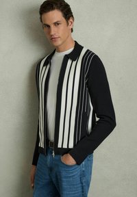 Jachetă cu fermoar, cu dungi negre și albe, cu guler, asociată cu o cămașă albă și blugi. Textură de material neted și croială slim.