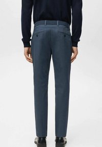 Man wearing dark blue slim-fit trousers, black belt, black shoes, and a dark long-sleeve top, standing facing away on a plain background.