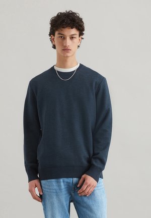 Pull à col rond bleu marine en tissu texturé, avec des poignets et un ourlet côtelés, porté par-dessus une chemise blanche, agrémenté d'une chaîne en argent.