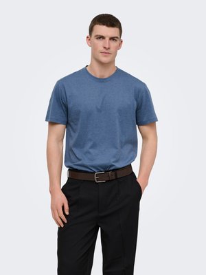 Young man wearing a blue short-sleeve t-shirt tucked into black pants with a brown belt, standing with one hand in pocket.