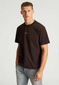 Bruine T-shirt met korte mouwen van katoen, voorzien van een ronde hals en minimale branding op de borst, gedragen met lichtblauwe jeans.