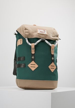 Sac à dos à rabat roulé vert et beige avec des bretelles en cuir, un cordon blanc et une étiquette de marque cousue à l'avant, posé sur un piédestal blanc.