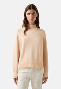 Vrouw met lang bruin haar, die een lichtbeige coltrui en crèmekleurige hoge taillebroek draagt, tegen een effen achtergrond.