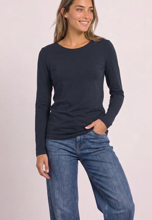 Kvinde smiler iført en navyblå langærmet skjorte og blå jeans, med den ene hånd i lommen, mod en ensfarvet lyse baggrund.