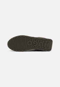Suela de un zapato en verde oliva con patrones de relieve pronunciados y el logotipo "HUGO" embellecido en la zona del talón, con un diseño grueso y texturizado.