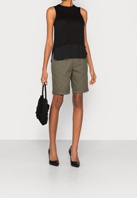Černý top bez rukávů s vrstveným lemem, olivově zelené šortky, černé vysoké podpatky a strukturovaná černá kabelka. Jednoduchá, neformální kombinace oblečení.