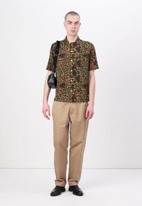 Camisa de print de leopardo en verde y marrón de manga corta hecha de una tela ligera, combinada con pantalones beige y zapatos negros. Bolsa de hombro negra.