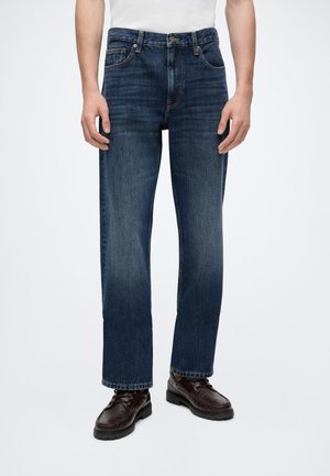 Mann trägt dunkelblaue Jeans mit geradem Bein und dunkelbraune Lederschuhe, steht vor einem schlichten hellen Hintergrund.