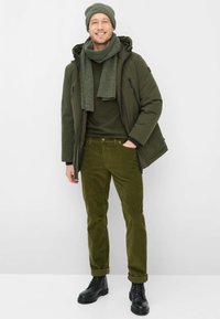 Groene gewatteerde jas met capuchon, bijpassende sjaal en muts, gecombineerd met ribfluwelen broek en zwarte veterlaarzen. Soepele texturen, samenhangend kleurenpalet.