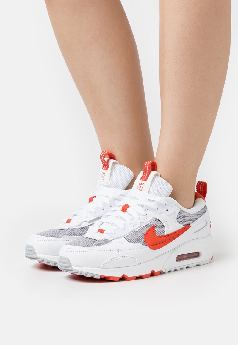 Jambes portant des baskets Nike Air Max blanches et grises avec un swoosh orange vif, des accents et des languettes sur fond blanc.