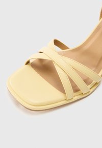 Sandale jaune à bout ouvert avec un devant carré et des brides croisées en cuir sur une semelle intérieure beige sur fond clair.
