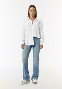 Witte, langermige shirt met knoopsluiting, geribbelde textuur. Gekleed met lichtblauwe flared jeans en witte sneakers. Eenvoudige, casual outfit.