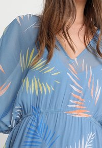 Mujer con un vestido azul de escote en V con patrones de hojas tropicales multicolores y una tela suelta y fluida.