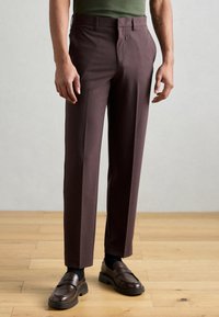 Calças castanho-escuras com corte ajustado, apresentando um franzido frontal plano, bolsos laterais e uma textura suave. Usadas com mocassins castanhos.