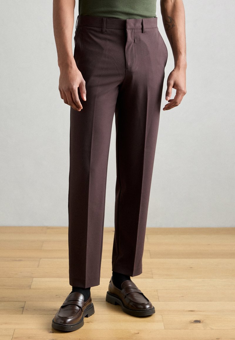 Calças castanho-escuras com corte ajustado, apresentando um franzido frontal plano, bolsos laterais e uma textura suave. Usadas com mocassins castanhos.