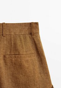 Brown linen shorts feature a flat waistband, pleated front, and a back pocket. The fabric has a textured finish, adding visual interest.